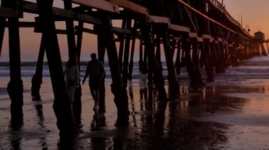 San Clementa California 'daki ahşap iskelenin altında güneş ve ışık destansı ışınlarıyla birlikte turuncu sahne pembe günbatımında el ele yürüyen çift.