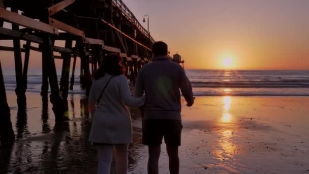 Couple walking away holding hands together at Scenic orange pink Sunset with epic rays of light and sun flare wooden pier in San Clementa California — Stock Video
