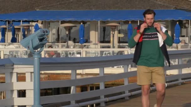 Красивий, молодий хлопець, чоловік у Томмі сорочці, шорти і окуляри ходять по ходьбі на hte pier — стокове відео