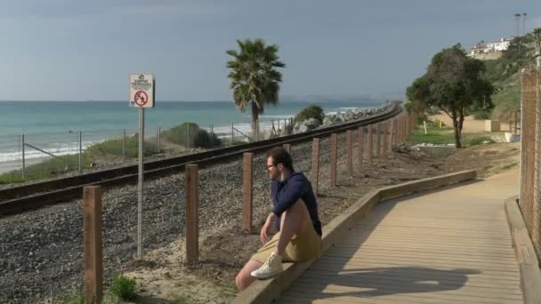 Un ragazzo bello e giovane, un uomo con una camicia blu navy, pantaloncini e occhiali sta camminando lungo la costa della California nel parco di San Clemente Linda Lane. Basso angolo attraverso le piante verdi — Video Stock