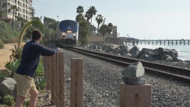 Un bel ragazzo giovane, un uomo con una camicia blu navy, pantaloncini che guardano il treno California Coast San Clemente Linda Lane Park . — Video Stock