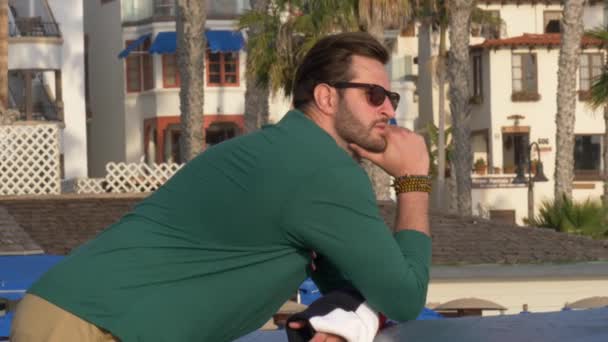 カリフォルニア海岸のサンクレメント・リンダ・レーン公園で見ているハンサムな若い男、ネイビーブルーのシャツ、ショートパンツ、眼鏡の男。緑の植物を通して低角度 — ストック動画