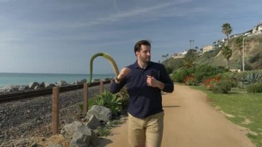 Yakışıklı, genç bir adam, lacivert gömlekli, şortlu ve gözlüklü bir adam California sahilinde yürüyor..