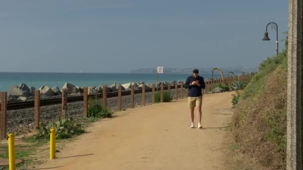 Egy jóképű, fiatal srác, egy férfi tengerészkék ingben, rövidnadrágban és szemüvegben sétál Kalifornia partjainál San Clemente Linda Lane parkban.. — Stock videók