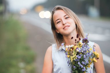 Genç sarışın bir kız elinde bir buket kır çiçeğiyle yolun arka planında kenarda dikiliyor. Kız çenesini kaldırdı ve gülümseyerek kameraya baktı..