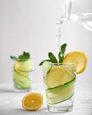 Kavramsal su, taze salatalık ve limon dilimleriyle dolu bir bardağa dökülür. Nane şekerli, açık beton arka planda, yüksek bir anahtarda.