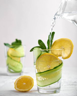 Kavramsal su, taze salatalık ve limon dilimleriyle dolu bir bardağa dökülür. Nane şekerli, açık beton arka planda, yüksek bir anahtarda.