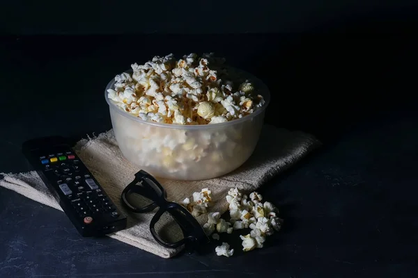 Skål med saltad popcorn, en TV-fjärrkontroll och glasögon på ett svart bord, selektivt fokus — Stockfoto