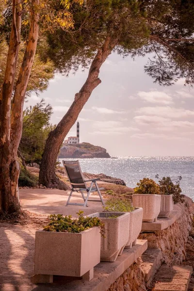 Farol sobre rochas à distância com espreguiçadeira em primeiro plano rodeado por árvores e flores, Portocolom, Maiorca, Espanha . — Fotografia de Stock