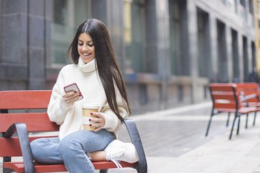 Modern ve gülümseyen genç kız elinde bir fincan kahveyle caddedeki bankta oturuyor.