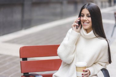 Modern ve gülümseyen genç bir kız elinde bir fincan kahveyle caddedeki bankta oturuyor.