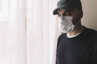 Hastalık ya da salgın hastalık için karantinadayken ev penceresinin yanında maske takan bir adam. Grip ve koronarivus kavramları