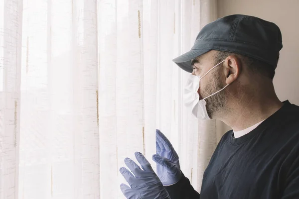 Man with face mask looking out the window of his home while quarantining for illness. Flu, coronarivus or pandemic concepts