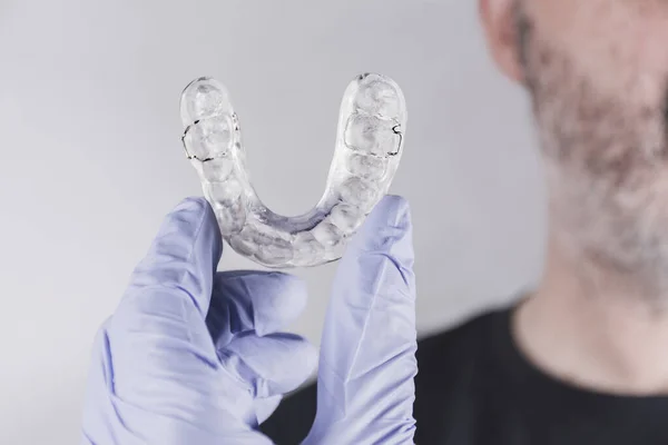 Hand with blue glove holding a dental splint, facing an adult man in front of white background