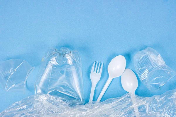 Top view of crushed plastic spoons, forks, bottles and cups as a disposable waste with copy space on bright blue background. Environmental pollution and litter recycling concept.