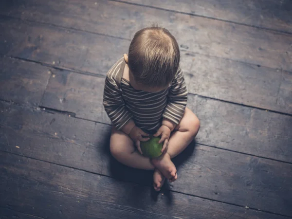 Little baby sitting on floorboards with apple — Stock Photo, Image