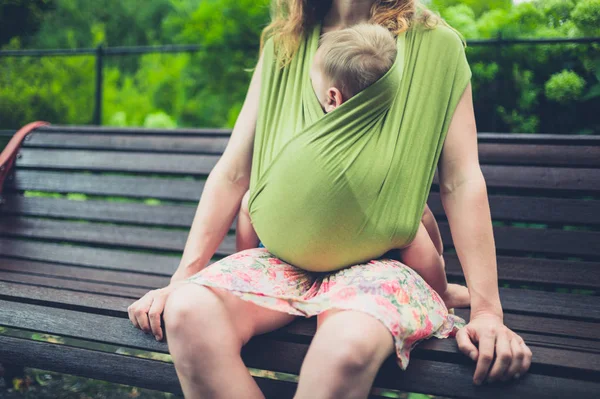 Мать с ребенком в стропе отдыхает на скамейке в парке — стоковое фото