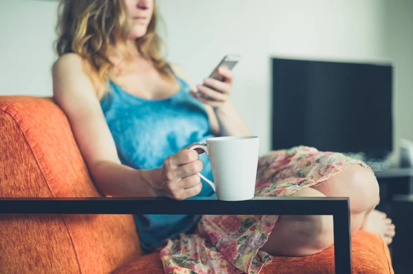 Mujer joven con taza y teléfono inteligente en casa —  Fotos de Stock