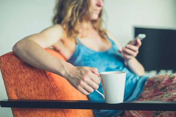 Jonge vrouw met cup en slimme telefoon thuis — Stockfoto