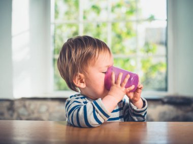 Sevimli küçük bir çocuk bir bardaktan içmeye masada oturuyor