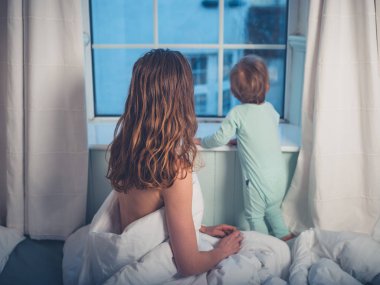 Genç bir anne ve küçük oğlu yatakta oturuyor ve sabah pencereden dışarı bakarak