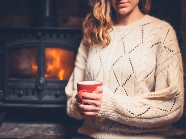 A young woman in a white jumper is sitting by the fireplace drinking from a mug