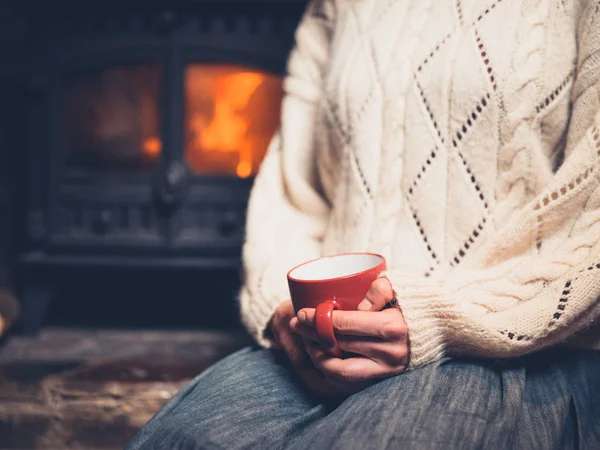 A young woman in a white jumper is sitting by the fireplace drinking from a mug