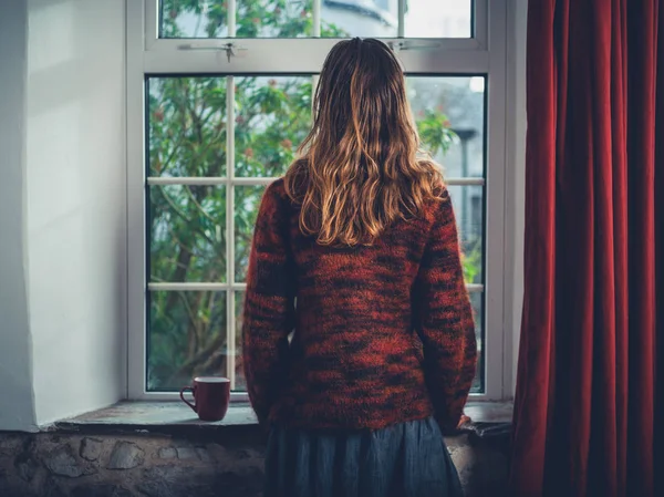 A young woman is standing by the window with a cup of tea