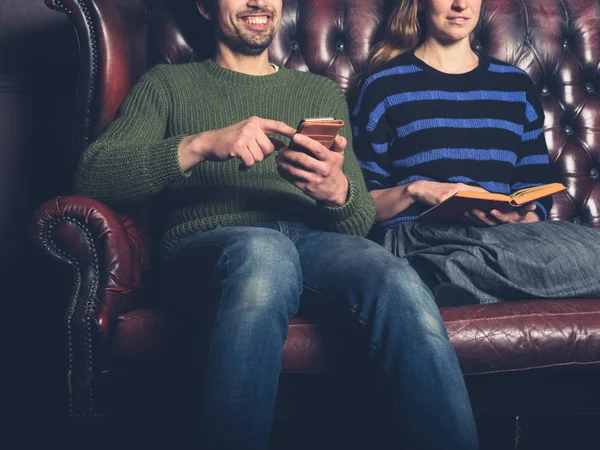 A happy young man on a sofa is using his smartphone next to a woman reading a book
