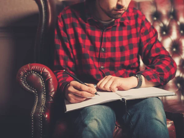 スマートな腕時計を身に着けている若い男は革のソファに座っているとノートに書いています — ストック写真