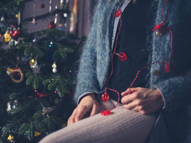 Genç bir kadın Noel ağacının yanında oturuyor. Elinde kalp şeklinde ışıklar var.