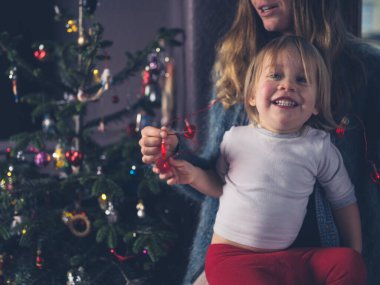 Bir anne ve bebeği Noel ağacının yanında oturmuş kalp şeklindeki ışıklara bakıyorlar.