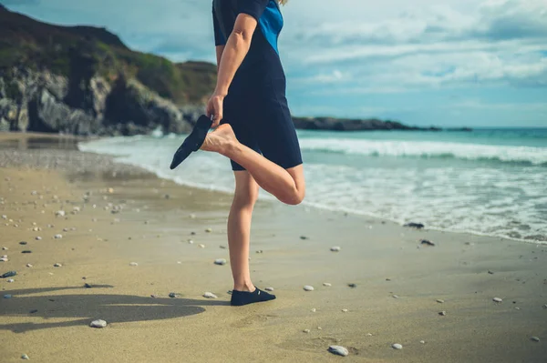 A young woman in a wetsuit on the beach is putting on her waterproof shoes