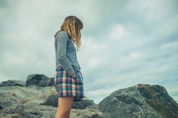 一个双手插在口袋里的年轻女子正站在多云的天空下的岩石上 — 图库照片