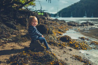 Küçük bebek sonbaharda nehrin kıyısındaki kayada oturuyor.