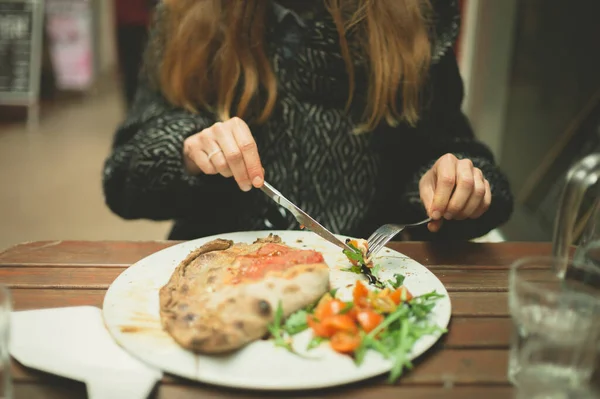 一位穿着冬衣的年轻女子在一家拱廊的室外餐桌边吃着一份带花生酱的披萨 — 图库照片