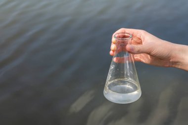 hand holding test tube for analyses with water on the background of the reservoir, the concept of water purity, pollution of water bodies, checking the quality of drinking water in cities, environmental