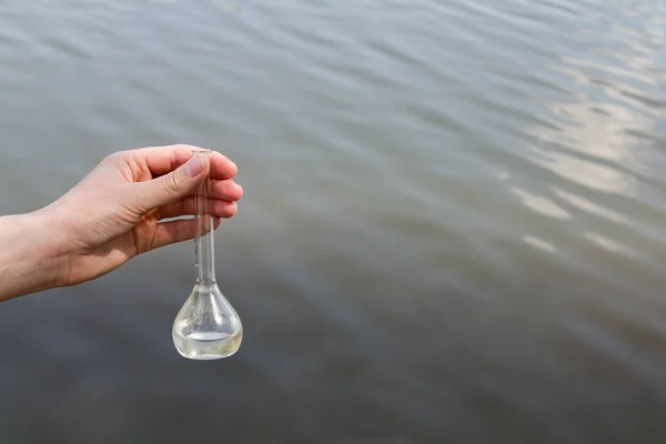 hand holding test tube for analyses with water on the background of the reservoir, the concept of water purity, pollution of water bodies, checking the quality of drinking water in cities, environmental