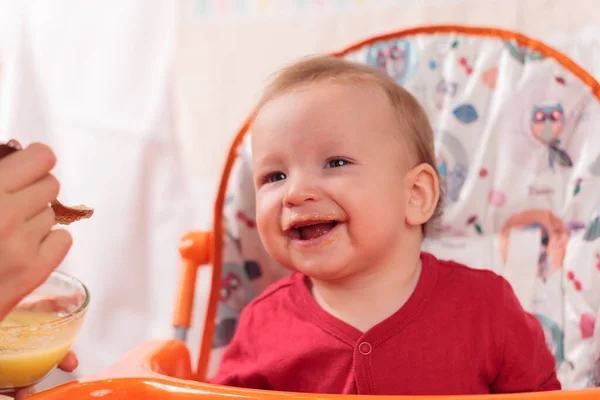 a small child eats with a spoon and sits and holds the edge of the feeding table in the nursery, grimy and stained with food, baby food concept, complementary foods, the first spoon