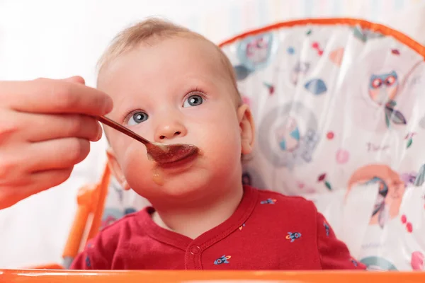 a small child eats with a spoon and sits and holds the edge of the feeding table in the nursery, grimy and stained with food, baby food concept, complementary foods, the first spoon