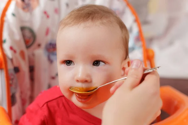 a small child eats with a spoon and sits and holds the edge of the feeding table in the nursery, grimy and stained with food, baby food concept, complementary foods, the first spoon