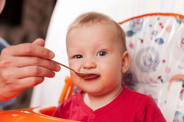 a small child eats with a spoon and sits and holds the edge of the feeding table in the nursery, grimy and stained with food, baby food concept, complementary foods, the first spoon