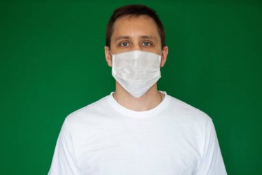 Tıbbi eldivenli bir adam yeşil arka planda beyaz bir tıbbi maske tutuyor.