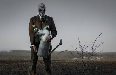 Elinde su bidonu olan gaz maskeli bir adam tek bir çiçekli ağacın yanında boş bir zeminde duruyor.