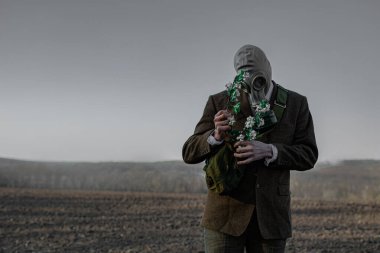 Elinde su bidonu olan gaz maskeli bir adam tek bir çiçekli ağacın yanında boş bir zeminde duruyor.