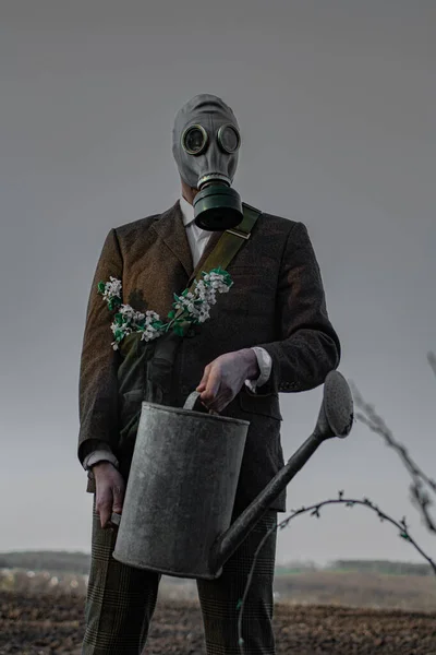 Ein Mann in einer Gasmaske mit einer Gießkanne in der Hand steht auf freiem Boden neben einem einzigen blühenden Baum — Stockbild Ein Mann Einer Gasmaske Mit Einer Gießkanne Der Hand Steht — Stockfoto
