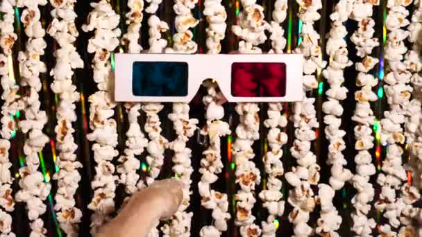 Katzenpfote spielt mit 3D-Brille. Defokussierter dynamischer Hintergrund von Girlanden aus Popcorn und goldglitzerndem Lametta. Komische Nachrichten, lustige Unterhaltung, Spaß. 4K-Video
