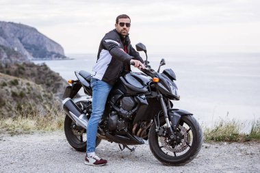 Young man biker with his black motorbike ready to drive, in front of the sea