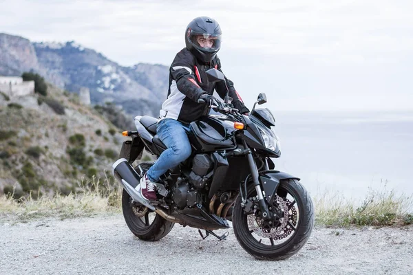 Pemuda pengendara motor dengan sepeda motor hitam siap untuk mengemudi, di depan laut — Stok Foto