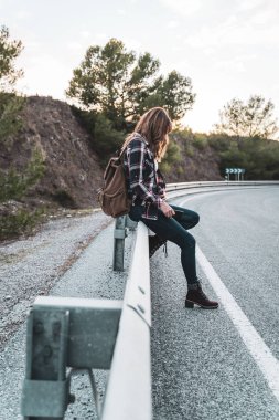 Young hip woman with her backpack hitchhiking on the road waiting for some car. Concept of exploration and adventures.